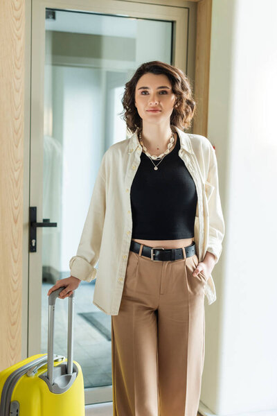 young woman in stylish casual clothes, white shirt, black crop top and beige pants standing with hand in pocket near yellow travel bag and glass door of contemporary hotel, travel lifestyle