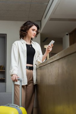 Beyaz gömlekli, siyah bluzlu ve bej pantolonlu genç ve şık bir kadın modern otelin lobisindeki resepsiyon masasının yanında sarı seyahat çantasıyla dikilirken cep telefonuyla mesajlaşıyor.