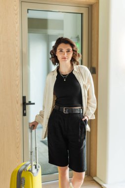 Modern otelin lobisindeki kameraya bakarken şık ve kendine güvenen beyaz gömlekli, kırışık bluzlu ve siyah şortlu kadın sarı bavulun ve cam kapının yanında el ele duruyor.