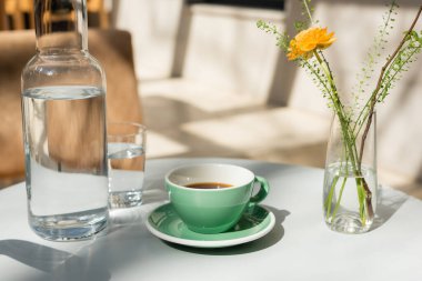 vase with yellow rose and green plants, glass and decanter with fresh pure water, saucer, cup with black coffee on white round table in morning sunlight, outdoor terrace of hotel cafe clipart