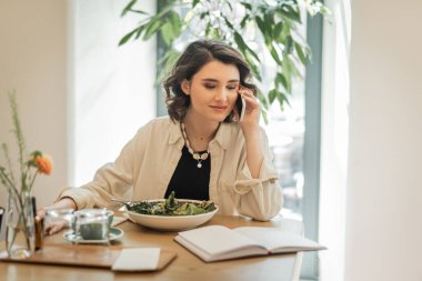 Moda kıyafetleri içindeki pozitif kadın cep telefonuyla konuşurken otel lobisinde taze sebze salatası, bardaklar ve bir fincan aromatik kahve ve iş hayatı entegrasyonunun yanında deftere bakıyor.