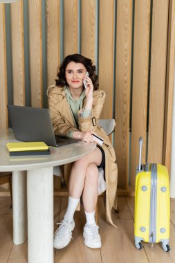 smiling woman in beige trench coat, with wavy brunette hair and tattoo talking on smartphone and holding credit card near laptop, notebooks on table and yellow suitcase in lobby of modern hotel clipart