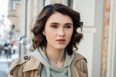 portrait of charming and fashionable woman with short brunette hair, in beige trench coat and grey hoodie looking away in European city on blurred background, street photography clipart