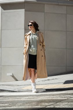 full length of trendy woman with wavy brunette hair crossing road in beige trench coat, grey hoodie, black shorts, white sneakers and dark sunglasses near modern building in city clipart