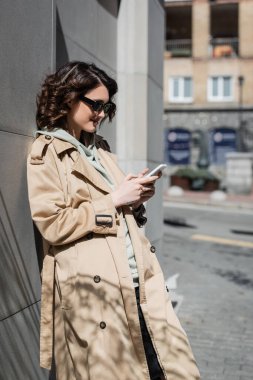 Kahverengi saçlı ve dövmeli, koyu güneş gözlüklü ve bej renkli trençkotlu neşeli ve modaya uygun bir kadın gri binanın yanında duruyor ve Avrupa 'da bir sokakta cep telefonuyla interneti tarıyor.