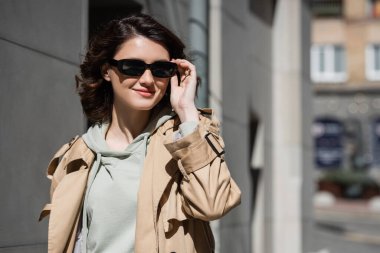 young and pleased woman with wavy brunette hair, in grey hoodie and beige trench coat adjusting dark stylish sunglasses and looking away on urban street on blurred background clipart