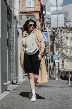 full length of fashionable and confident woman in beige trench coat, grey hoodie, black shorts, white sneakers and dark sunglasses walking on street in European city and looking away clipart