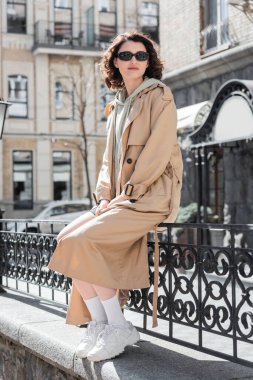 Moda kıyafetleri içinde, bej rengi trençkotlu, beyaz spor ayakkabılı, siyah gözlüklü sahte çitlerde oturan ve Avrupa şehrinin caddesine bakan modaya uygun genç bir kadın.