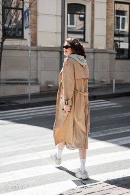 full length of fashionable woman in grey hoodie, beige trench coat and white sneakers crossing road and looking away in European city, travel lifestyle, urban fashion  clipart