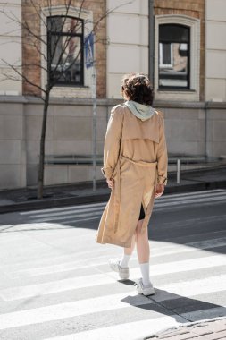 full length of young woman with wavy brunette hair, in beige trench coat, grey hoodie and white sneakers crossing road on sunny street of European city, back view, urban fashion, travel lifestyle  clipart