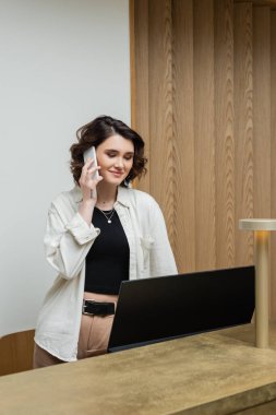Mutlu genç resepsiyonist, dalgalı esmer saçlı, dövmeli, şık kıyafetleri olan, cep telefonuyla konuşan otel lobisindeki bilgisayar monitörü ve lambasının yanında çalışan.