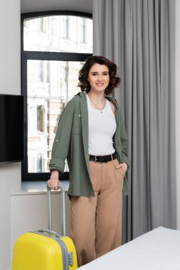 Dalgalı esmer saçlı, şık giyinmiş gülümseyen kadın pencerenin yanında el ele dururken kameraya bakıyor. Gri perdeler, lcd TV ve rahat otel odasında yatak.