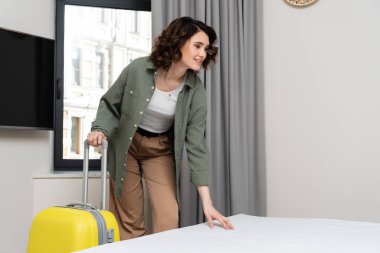 Şık giyinmiş neşeli bir kadın, dalgalı esmer saçlı sarı valiz, pencere yakınındaki otel odasında rahat bir yatağa dokunuyor, gri perdeler ve boş ekranlı lcd TV, seyahat yaşam tarzı.