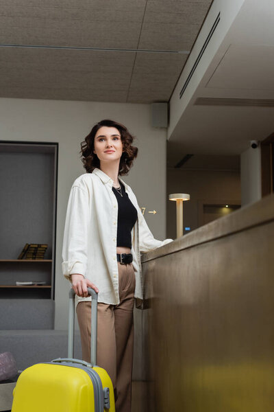 young and trendy woman with short brunette hair, in white shirt, black crop top and beige pants standing with yellow suitcase near reception desk and looking away in lobby of modern hotel