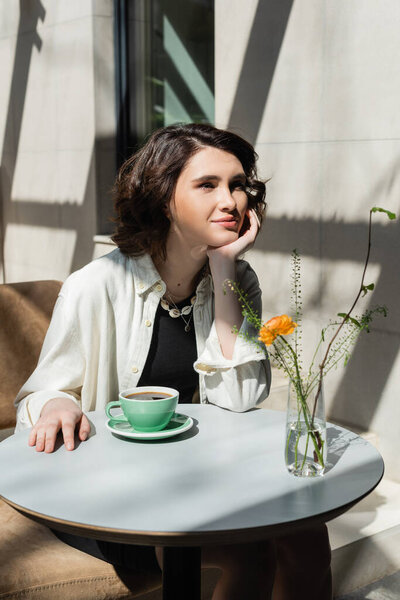 Улыбающаяся женщина сидит за белым круглым столом возле чашки черного ароматного кофе, блюдца, стеклянной вазы с желтой розой и зелеными растениями на летней террасе гостиничного кафе при утреннем солнечном свете