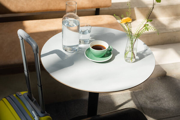 чемодан возле белого круглого стола с чашкой черного ароматного кофе, блюдцем, графином и стаканом пресной чистой воды, вазой с зелеными растениями и желтой розой на открытой террасе гостиничного кафе