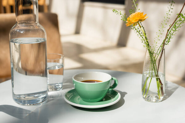ваза с желтой розой и зелеными растениями, стекло и графин со свежей чистой водой, блюдце, чашка с черным кофе на белом круглом столе в утреннем солнечном свете, открытая терраса кафе отеля