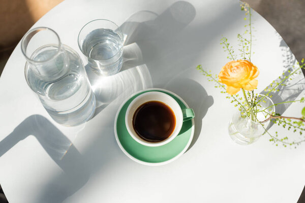 верхний вид стекла и графина с чистой водой, чашка с черным кофе, блюдце, ваза с желтой розой и зелеными растениями на белом круглом столе в утреннем солнечном свете, кафе отеля, летняя терраса