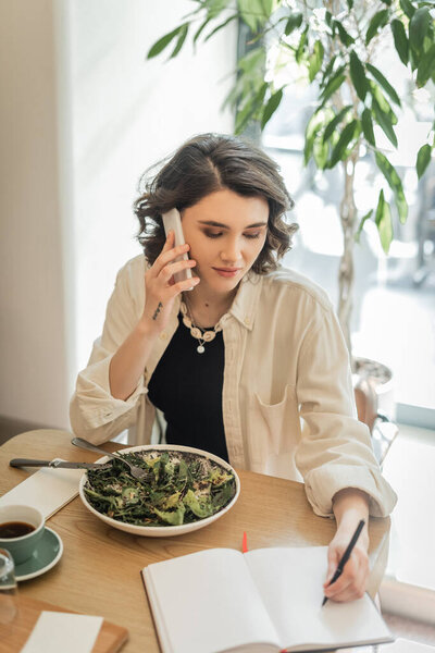 young tattooed woman with short brunette hair talking on smartphone and writing in notebook near vegetable salad and cup of aromatic coffee on table in lobby cafe of modern hotel, work and travel