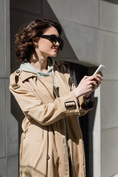 快乐的女人 戴着深色时髦太阳镜 穿着米黄色的风衣 留着波浪般的黑发 纹身在城市街道阳光下的灰色大楼附近的智能手机上聊天 旅行生活方式 — 图库照片
