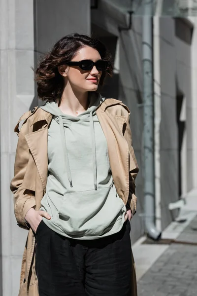 stock image confident young woman with wavy brunette hair, in dark stylish sunglasses, grey hoodie and beige trench coat standing with hands in pockets of black shorts and looking away on street