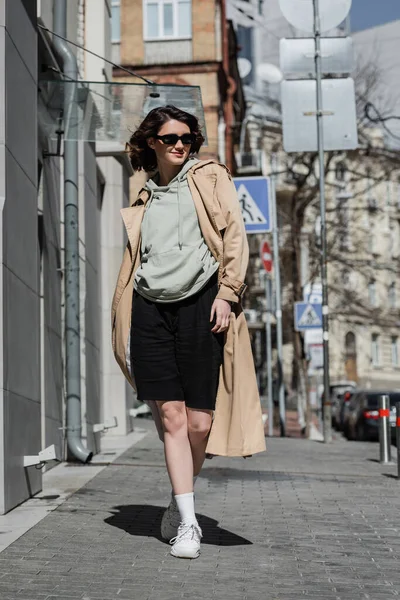 stock image full length of fashionable and confident woman in beige trench coat, grey hoodie, black shorts, white sneakers and dark sunglasses walking on street in European city and looking away