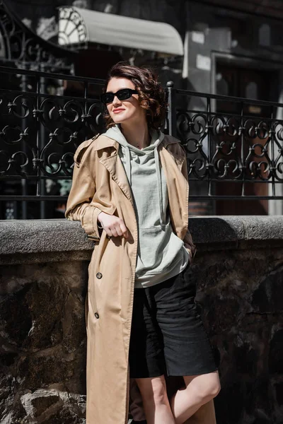 stock image travel lifestyle, trendy and cheerful woman in grey hoodie, beige trench coat and stylish sunglasses standing with hand in pocket of black shorts and looking away near forged fence on city street