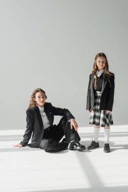 Çalışan anne ve çocuk, takım elbiseli iş kadını üniformalı bir kızın yanında oturuyor gri arka planda ekose etekli, ceketli, yeni okul yılındaki, kameraya bakan, resmi kıyafetli. 