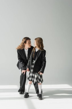 Çalışan anneye bakan liseli kız, okul üniformalı neşeli kız gri arka planda takım elbiseli iş kadınıyla duruyor, resmi kıyafetler, moda ailesi, kaynaşma, modern ebeveynlik. 