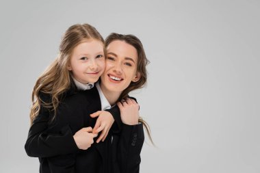 smiling schoolgirl hugging mother in suit, girl in school uniform and her mom in business attire on grey background, fashionable family, bonding, modern parenting, back to school  clipart