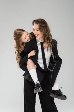 Anne-kız eğleniyor, neşeli kadın okul kızına biniyor, resmi kıyafetlerle çalışan anne ve çocuk gri arka planda stüdyoda, modern ebeveynlikte çalışıyor. 