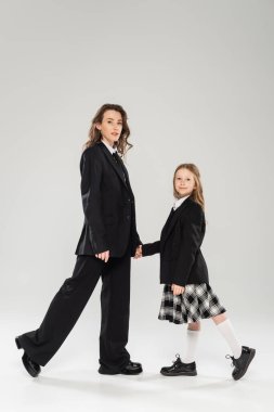 Anne ve okul kızı el ele tutuşuyor, iş elbiseli bir kadın ve okul üniformalı mutlu bir kız gri arka planda, modern ebeveynlikte, okula geri dönüyor. 