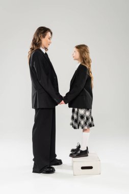 Anne ve çocuk el ele tutuşuyor, iş elbiseli mutlu bir kadın ve okul üniformalı bir çocuk gri arka planda bir taburede duruyor, modern ebeveynlik, yüz yüze.