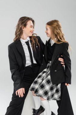 Çalışan anne kız, mutlu iş kadını üniformalı bir çocuğun yanında oturan gri arka planda stüdyo, resmi kıyafetler, modern ebeveynlik, moda çekimi. 