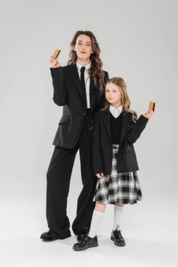 Moda annesi ve kızı, takım elbiseli iş kadını ve gri arka planda kredi kartı tutan okul kızı, modern ebeveynlik, finansal öğrenim, bütçe, para yönetimi  