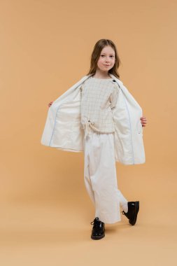 Beyaz takım elbiseli ve siyah ayakkabılı modaya uygun genç kız bej arka planda dururken kameraya bakıyor, şık kıyafet, resmi kıyafet, çocuk modeli, moda belirleyicisi, tarz, spor ceketi gösteriyor.