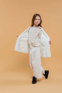 trendy preteen girl in white suit and black shoes looking at camera while standing on beige background, fashionable outfit, formal attire, child model, trendsetter, style, demonstrating blazer clipart