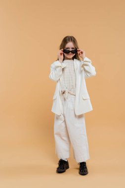 Beyaz takım elbiseli, siyah ayakkabılı, güneş gözlüğü takan, bej arka planda duran, modaya uygun giyinen, çocuk mankeni, moda belirleyicisi, moda belirleyicisi, stili olan genç kız. 