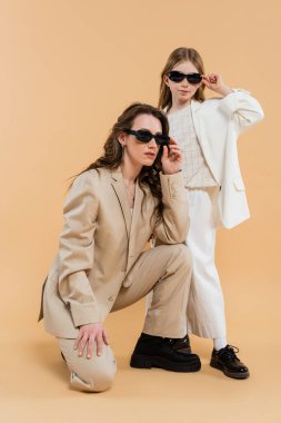 Modern aile, takım elbiseli ve güneş gözlüklü şık anne-kız, bej arka planda oturan iş kadını, şık kıyafetler, resmi kıyafetler, çalışan anne, moda belirleyici 