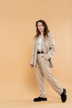 Moda trendi konsepti, dalgalı saçlı, şık takım elbiseli ve bej arka planda kameraya bakan genç bir kadın, el ele, klasik tarz, profesyonel kıyafet. 