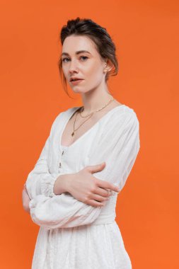 Yaz trendleri, güneş giysili manken kameraya bakıyor ve turuncu arka planda katlanmış kollarla duruyor, şık poz veriyor, beyaz giyimli bayan, canlı arka plan, moda, genç kadın. 