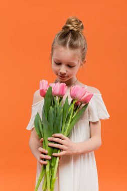 Yaz modası, elinde turuncu arka planda pembe laleler, moda ve tarz konsepti, çiçek buketi, şık çocuk, canlı renkler tutan beyaz güneş elbisesi içindeki genç kız. 