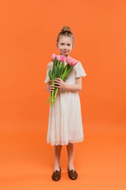 Yaz modası, turuncu arka planda pembe laleler tutan mutlu genç kız elbisesi, moda ve tarz konsepti, bir buket çiçek, şık çocuk, canlı renkler, tam boy 