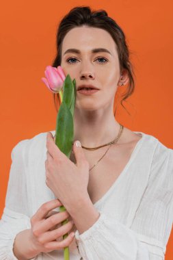 Günlük moda, elinde pembe lale tutan ve turuncu arka planda duran beyaz elbiseli genç kadın, beyaz, canlı arka plan, moda ve doğa, yaz, portre 