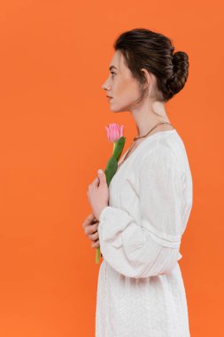 Pembe lale tutan ve turuncu arka planda duran beyaz elbiseli genç bir kadının yan görüntüsü. Beyaz, canlı arka plan, moda, yaz, portre, şehvet, günlük moda. 