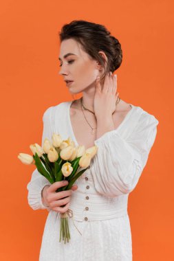 Moda konsepti, elinde sarı lale tutan, turuncu arka planda duran beyaz elbiseli genç kadın, canlı arka plan, moda, yaz, bir buket çiçek. 