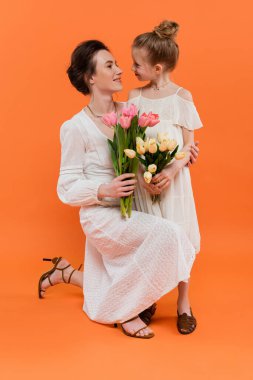 Çiçekli mutlu anne ve kız, lale tutan genç kadın ve kız ve kız turuncu arka planda poz veriyorlar, yaz modası, güneş elbiseleri, kadın dayanışması 