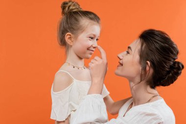 Aile bağları, neşeli anne turuncu arka planda reşit olmayan kızın burnuna dokunuyor, beyaz güneş elbiseleri, modern ebeveynlik, yaz modası, birliktelik, aşk, moda aile. 