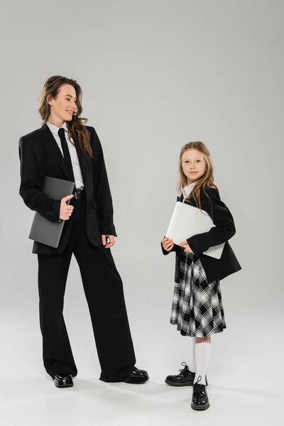mother and daughter, digital nomadism, remote work, e learning, happy businesswoman in suit and schoolgirl standing together with laptops on grey background in studio, modern parenting 