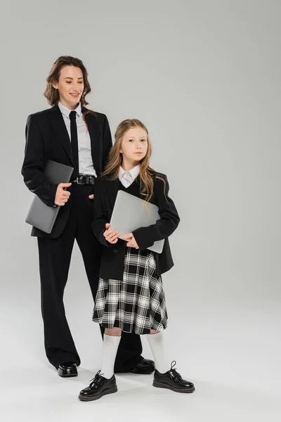 stock image happy mother and daughter, digital nomadism, remote work, e learning, businesswoman in suit and schoolgirl standing together with laptops on grey background in studio, modern parenting 